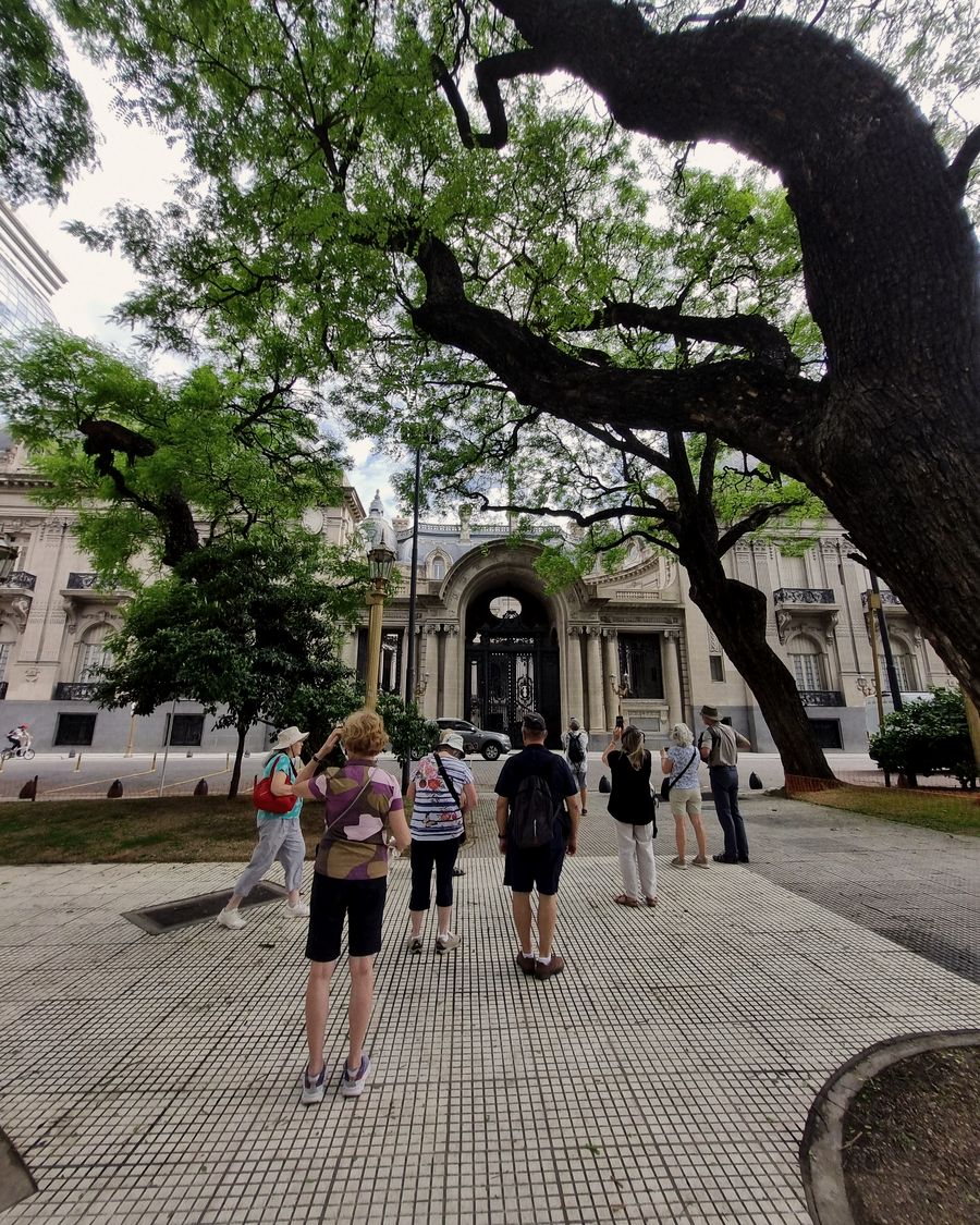 Small group of travelers looking toward the entrance of Palacio San Martín in Retiro, Buenos Aires, shaded by a jacaranda tree